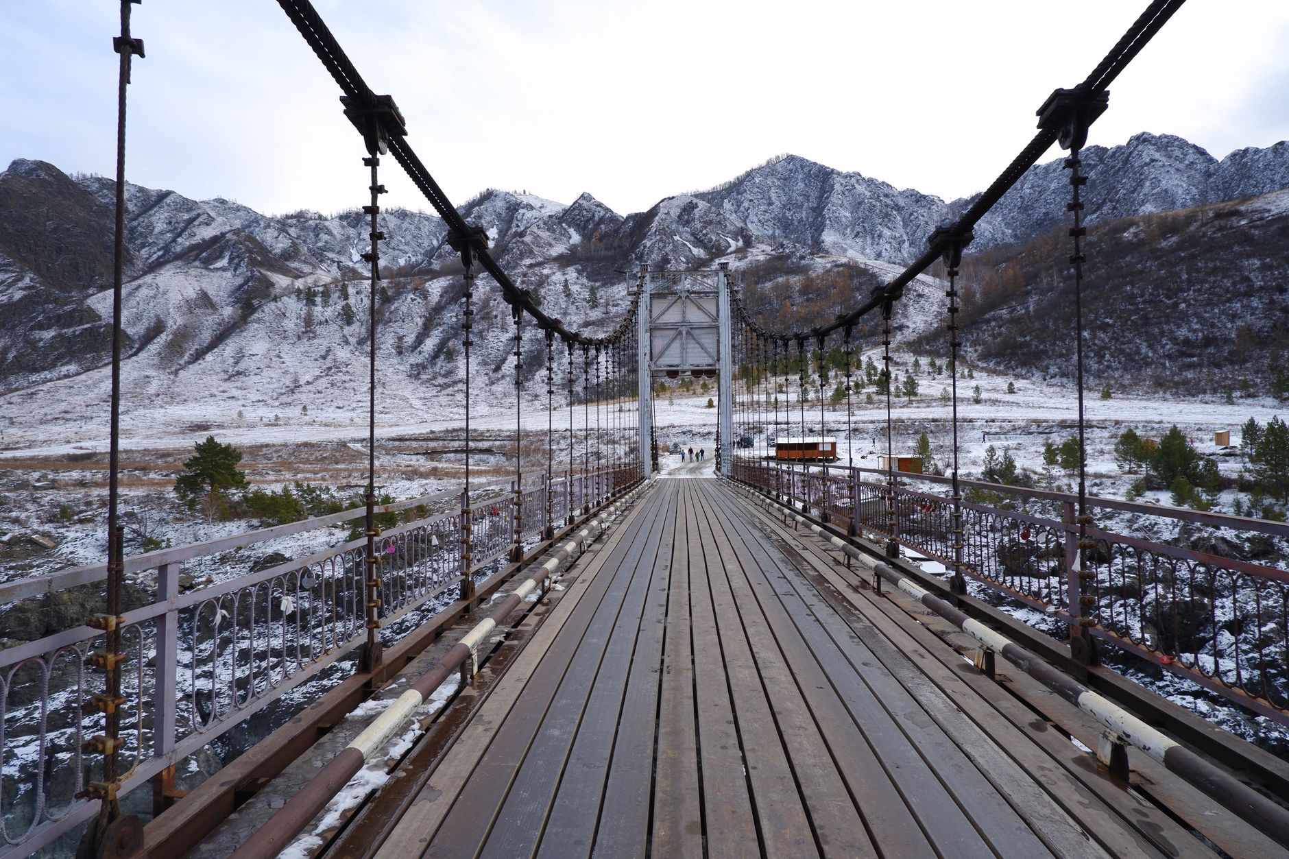 Narrow wooden suspension bridge crossing a glacial river with towering mountain peaks in the background